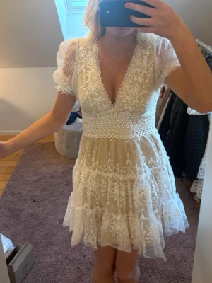 Vit spetsklänning med volanger - Romantisk kort klänning i vit spets med v-ringning och puffärmar. Klänningen har fina broderade blommor, markerad midja med spetsdetalj och volanger i kjolen. Perfekt för sommarens alla tillfällen.