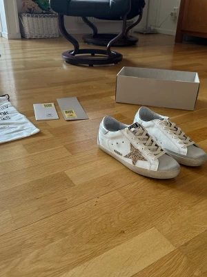 Golden Goose vita sneakers med glitterstjärna - Säljer ett par vita Golden Goose sneakers med en stor glittrig guldstjärna på sidan och silverdetalj vid hälen. Skorna har beige snören och klassisk låg profil. Perfekta för dig som vill sticka ut med coola detaljer och premiumkänsla.