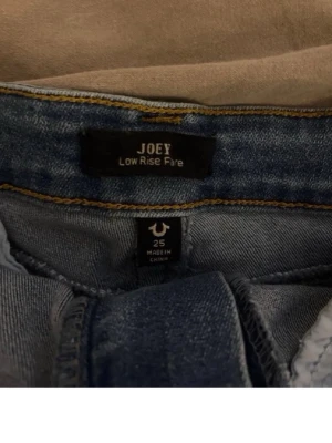 True Religion Joey Low Rise flare jeans - Säljer ett par blå Joey Low Rise flare jeans från True Religion. De har utsvängda ben, låg midja och snygga detaljer på bakfickorna med knappar. Materialet är klassisk denim och passformen är riktigt cool och trendig.