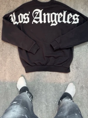 Svart sweatshirt Los Angeles Black Squad - Cool svart oversized sweatshirt från Black Squad i storlek XS. Stor vit text 'Los Angeles' på ryggen i gotisk stil. Mjuk insida och ribbade muddar vid ärmar och nederkant. Perfekt för en avslappnad streetwear-look.