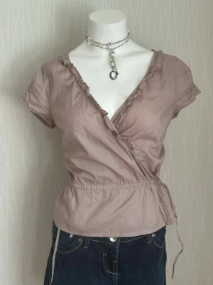 Beige omlottopp med volangdetalj - Supersöt beige omlottopp med korta ärmar och volangkant längs v-ringningen. Toppen har knyt i sidan och resår i midjan för en snygg passform. Perfekt att styla med jeans eller kjol för en trendig look.