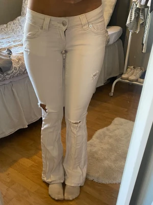 Vita bootcut jeans med slitningar - Snygga vita bootcut jeans med slitna detaljer på benen och klassiska fem fickor. Jeansen har låg midja och lätt utsvängda ben, perfekt för en trendig look. Materialet är denim med stretch för extra komfort.