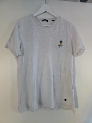 Vit t-shirt från Jack & Jones - Vit t-shirt från Jack & Jones med ett litet ananastryck och texten 'Premium Summer' på bröstet. Klassisk rund hals och korta ärmar. Materialet är mjuk bomull och t-shirten har en avslappnad passform. Perfekt för dig som gillar enkel stil med en twist.