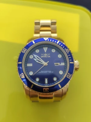 Guldfärgad klocka från Invicta - Snygg guldfärgad klocka från Invicta med blå urtavla och blå vridring. Klockan har tydliga indexmarkeringar, datumfönster och robust länkarmband i metall. Perfekt accessoar för dig som vill sticka ut med stilren design och lyxig känsla.