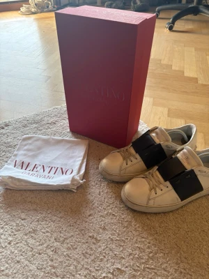 Valentino Garavani Open  - Säljer ett par vita sneakers från Valentino Garavani med bred mörkblå detalj över snörningen. Skorna är tillverkade i skinn och har klassisk rund tå och platt sula. Medföljer originalkartong och dustbag. Perfekta för dig som vill ha en lyxig och stilren look.