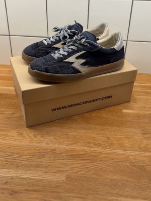 MOA Concept blåa sneakers - Snygga sneakers från MOA Concept i mörkblå mocka med beige detaljer och en cool blixtlogga på sidan. Skorna har vita och gråa snören samt en brun gummisula. Perfekta för dig som vill ha en unik och trendig look.