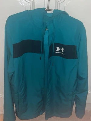 vindjacka från Under Armour - Snygg vindjacka från Under Armour med svart panel över bröstet och vit logga. Jackan har huva med dragsko, hel dragkedja framtill och två sidofickor. Perfekt för blåsiga dagar och sportiga outfits.