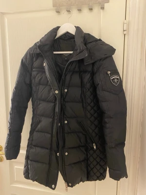 Svart lång dunjacka från Rockandblue - Snygg svart dunjacka från Rockandblue med quiltade detaljer på sidorna och ärmen. Jackan har hög krage, dragkedja och knappar framtill samt flera fickor med dragkedja. Märkespatch på ena ärmen och figurnära passform. Köpt för några år sedan för 3000 kr.