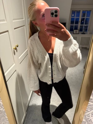 Vit stickad kofta med pärlknappar - Säljer en vit stickad kofta med v-ringning och pärlknappar framtill. Koftan har ballongärmar och ribbade muddar, vilket ger en avslappnad och trendig look. Perfekt att slänga över ett linne eller t-shirt för att höja stilen. Storlek L men skulle säga mer S/M