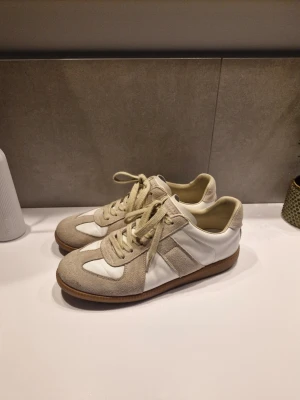 Maison margiela skor vita - Snygga sneakers i vitt och beige med klassisk design. Skorna har paneler i mocka och släta partier i läder, samt beige snörning och en brun gummisula. Perfekt för dig som gillar stilrena och tidlösa sneakers.