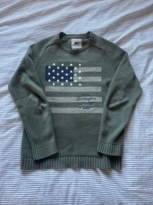 Grön stickad tröja från Lexington - Snygg grön stickad tröja från Lexington med amerikansk flagga i blått, vitt och grått på bröstet. Tröjan har rund halsringning, ribbade muddar och är tillverkad i mjukt material. Perfekt för dig som gillar klassisk och avslappnad stil.
