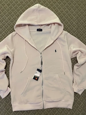 Rosa hoodie från Polo Ralph Lauren - Snygg ljusrosa hoodie från Polo Ralph Lauren med dragkedja, två stora fickor framtill och klassisk huva med dragsko. Ikoniska loggan broderad på bröstet. Tillverkad i mjukt bomullsmaterial som känns skönt mot huden. Perfekt för en avslappnad och trendig look.
