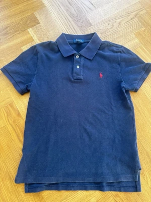 Marinblå pikétröja från Ralph Lauren - Klassisk marinblå pikétröja från Ralph Lauren med korta ärmar och krage. Tröjan har knappar framtill och den ikoniska röda loggan broderad på bröstet. Perfekt för en stilren och avslappnad look. 