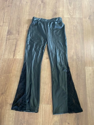 Svarta utsvängda leggings med spets - Säljer ett par svarta leggings med utsvängda ben och snygga spetsdetaljer längs sidorna. Materialet är glansigt och stretchigt, vilket ger en cool look. Perfekta för dig som vill sticka ut lite extra med din outfit!