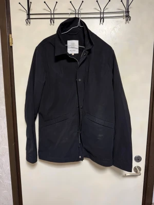 Svart jacka från Elvine - Stilren svart jacka från Elvine med klassisk krage och knäppning framtill. Jackan har två snedställda fickor och en minimalistisk design som passar perfekt till streetwear eller en mer dressad look. Materialet är slätt och lätt, perfekt för vår och höst.