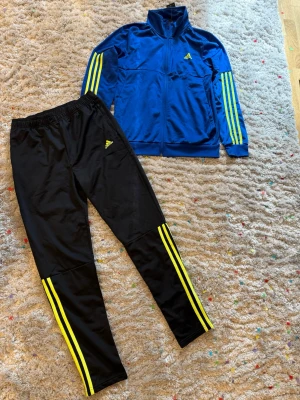 Adidas blå träningsset med gula ränder - Snyggt träningsset från Adidas med blå zip-jacka och svarta byxor. Båda plaggen har de klassiska gula Adidas-ränderna längs ärmar och ben samt Adidas-logga på bröst och lår. Perfekt för träning eller chill. Materialet är mjukt och stretchigt, troligen polyester , storlek 176 / XL för barn 