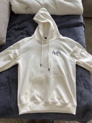 Vit hoodie från Amiri med logga - Säljer en stilren vit hoodie från Amiri med stor logga på ryggen och mindre logga på bröstet. Hoodien har klassisk känguruficka, ribbade muddar och dragsko i huvan. Perfekt för dig som gillar streetwear och vill ha något trendigt och bekvämt. Priset kan diskuteras. 