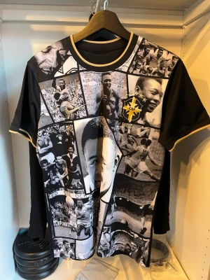 Svart t-shirt med Pelé collage - Unik svart t-shirt med collage av svartvita foton på Pelé och Brasilien-logga på framsidan. Baksidan har texten 'Pelé' och nummer 10 i vitt. T-shirten har korta ärmar och gulddetaljer vid hals och ärmslut. Perfekt för fotbollsfans!