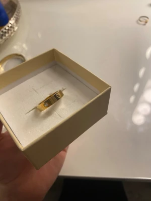 Guldpläterad ring med hjärta och stenar - Snygg Guldpläterad ring med platt design, dekorerad med små gnistrande stenar och ett ingraverat hjärta. Perfekt för dig som gillar stilrena accessoarer med en touch av romantik. Levereras i ask.