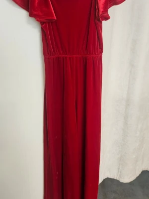 Röd sammetsjumpsuit med vida ben - Snygg röd jumpsuit i mjuk sammet med vida ben och korta, fladdriga ärmar. Resår i midjan för en bekväm passform och elegant look. Perfekt för dig som vill sticka ut med en trendig och färgstark outfit.