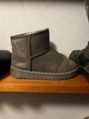 Gråa fluffiga tofflor i mocka-look - Mysiga gråa tofflor i mocka-liknande material med fluffigt foder och grov sula. Klassisk design med synliga sömmar och rundad tå. Perfekta för att hålla fötterna varma hemma eller när du snabbt ska ut. Märket är Fashion och storleken är 35.