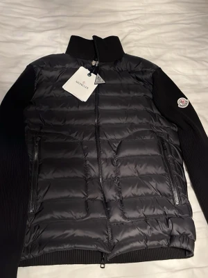 Cardigan från Moncler  - Snygg svart cardigan från Moncler med ribbstickade svarta ärmar och hög krage. Cardigan har två dragkedjeförsedda fickor framtill och klassisk Moncler-logga på ärmen. Materialet är en kombination av dun och stickat tyg för en modern look.