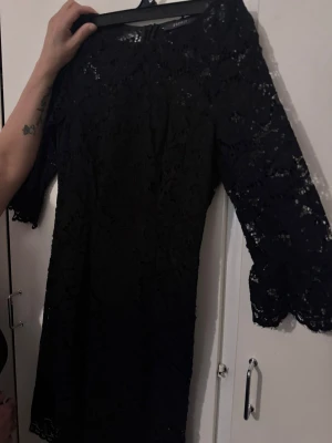Svart spetsklänning med trekvartsärm - Snygg svart klänning i spets med trekvartsärmar och rund halsringning. Klänningen har en elegant, figurnära passform och är helfodrad med transparenta spetsdetaljer på ärmarna. Perfekt för dig som gillar stilrena och klassiska plagg.