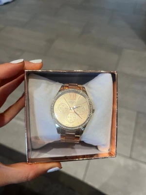Guldig Regal klocka med kristaller - Snygg rose klocka från Regal med metallarmband och glittrande kristaller runt urtavlan. Urtavlan har flera små visare och romerska siffror upptill. Perfekt accessoar för att lyfta din stil. Endast ett fåtal repor som syns på bilden därav priset 