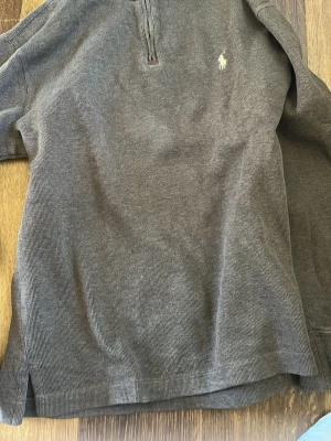 Grå ziptröja från Polo Ralph Lauren - Mysig grå ziptröja från Polo Ralph Lauren med liten broderad logga på bröstet. Tröjan har halv dragkedja framtill, ribbade muddar och är tillverkad i ett mjukt, strukturerat tyg. Perfekt för en avslappnad och stilren look.