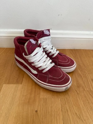 Vinröda Vans Sk8-Hi sneakers - Säljer ett par klassiska vinröda Vans Sk8-Hi sneakers med vit sula och vita snören. Skorna har den ikoniska vita Vans-sidan och är tillverkade i canvas med detaljer i mocka. Perfekta för dig som gillar streetwear och vill ha en tidlös look.
