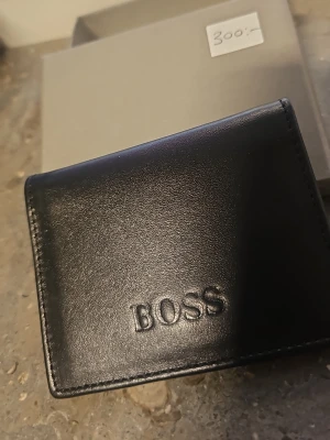 Svart plånbok från BOSS i läder - Stilren svart plånbok från BOSS i äkta läder med präglad logga på framsidan. Plånboken har flera kortfack och en tryckknapp för stängning. Perfekt för dig som vill ha en klassisk och snygg accessoar till dina kort.
