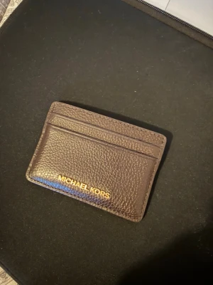 Korthållare - Stilrent kortfodral i mörkbrunt läder från Michael Kors med guldfärgad logotyp på framsidan. Fodralet har flera fack för kort och är tillverkat i ett strukturerat lädermaterial som känns lyxigt och modernt. Perfekt för dig som vill ha koll på dina kort och samtidigt hålla det snyggt.