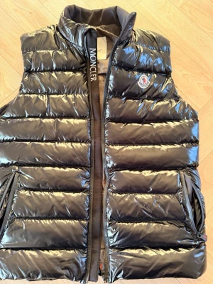 Svart dunväst från Moncler - Snygg svart dunväst från Moncler med glansig finish och klassisk quiltad design. Västen har hög krage, dragkedja framtill och två fickor med dragkedja. Moncler-logga på bröstet och broderad text längs dragkedjan. Perfekt för lager-på-lager-stil.