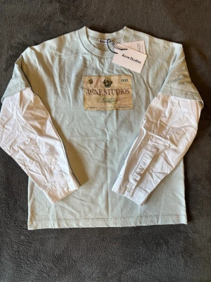 Ljusblå långärmad tröja Acne Studios - Ljusblå långärmad tröja från Acne Studios med tryckt vintageinspirerad etikett på bröstet. Tröjan har rund halsringning och är tillverkad i mjukt bomullsmaterial. Perfekt för en avslappnad och trendig look.