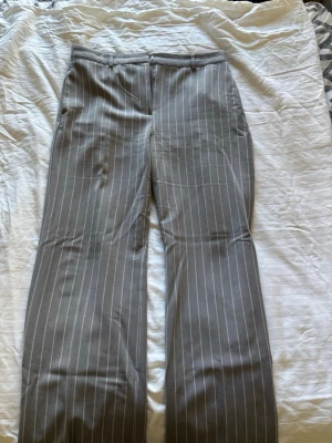 Grå kostymbyxor med vita ränder - Snygga grå kostymbyxor med vita kritstrecksränder och raka ben. Byxorna har klassisk passform, fickor och hällor för skärp. Perfekta för en stilren och trendig look. Materialet känns mjukt och följsamt, vilket gör dem bekväma att bära hela dagen.