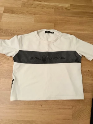 Vit Sail Racing t-shirt med svart detalj - Vit t-shirt från Sail Racing med en svart, mönstrad panel över bröstet och logga tryckt i svart. Rund halsringning och korta ärmar. Liten svart etikett med logga på vänster sida. Snygg och sportig stil, perfekt för en avslappnad look.