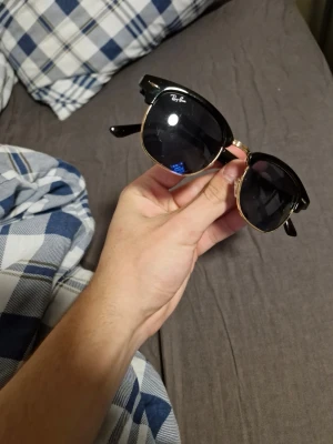 Svarta Ray-Ban solglasögon clubmaster - Snygga svarta Ray-Ban solglasögon i clubmaster-modell med guldfärgade detaljer. Klassisk design med svarta bågar och mörka glas. Perfekta för att lyfta din stil under soliga dagar.