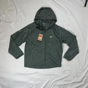 Grå Nike Miller Windbreaker - Snygg grå vindjacka från Nike med huva och dragkedja framtill. Jackan har en minimalistisk design med Nike-logga på bröstet och är tillverkad i lätt, vindtätt material. Perfekt för blåsiga dagar och sportiga outfits. Den sitter lite löst och luftigt passande för våren/sommaren. 