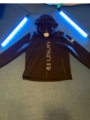 Svart vindjacka från Moncler - Snygg svart vindjacka från Moncler med stor logga vertikalt på framsidan. Jackan har huva, två dragkedjeförsedda fickor och justerbar nederkant. Tillverkad i lätt och slitstarkt material, perfekt för blåsiga dagar. Klassisk Moncler-patch på ärmen.