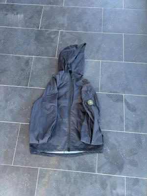 Svart vindjacka från Stone Island - Snygg svart vindjacka från Stone Island med huva och dragkedja framtill. Jackan har den klassiska Stone Island-loggan på vänster ärm och är tillverkad i ett lätt, vindtätt material. Perfekt för dig som vill ha en stilren och funktionell jacka.