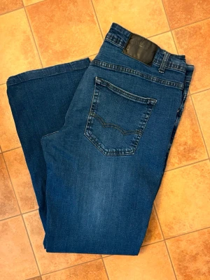 Blå bootcut jeans från Enzo W36 L32 - Snygga blå bootcut jeans från enzo med klassisk femficksdesign och subtila slitningar. Jeansen har normal midja, raka ben och är tillverkade i mjuk denim med stretch för extra komfort. Perfekta för en avslappnad och trendig look.