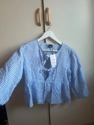 Blåvitrutig blus med knyt från Gina Tricot - Säljer min ljusblåa rutiga blus med två knyten där framme man kan välja att ha öppet eller knytet, blusen har trekvarts armar och är aldrig använd med prislappen kvar😊 (nypris 399) 