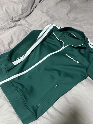 Grön track jacket från Palm Angels - Snygg grön track jacket från Palm Angels med vita ränder längs ärmarna och vit dragkedja. Jackan har ståkrage, fickor med dragkedja och logga broderad på bröstet. Perfekt för en sportig och trendig look.