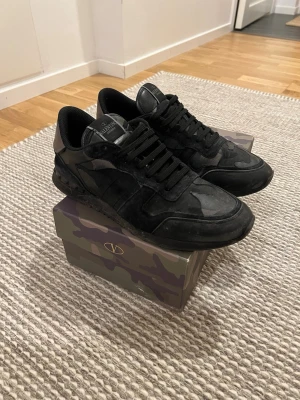 Svarta Valentino Garavani sneakers - Snygga svarta sneakers från Valentino Garavani med kamouflagemönster och detaljer i mocka och läder. Skorna har svarta skosnören, låg profil och en unik häl i metallic-brons. Perfekta för dig som gillar exklusiva och trendiga sneakers.