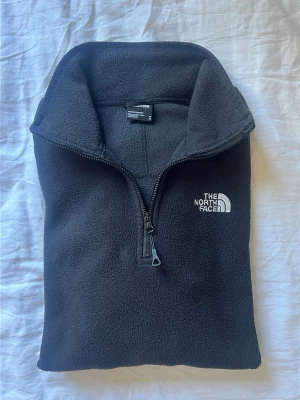 Svart fleece-tröja från The North Face - Mysig svart fleece-tröja från The North Face med halv dragkedja och hög krage. Tröjan har broderad logga på bröstet och är tillverkad i mjukt fleecematerial som håller dig varm under kyliga dagar.