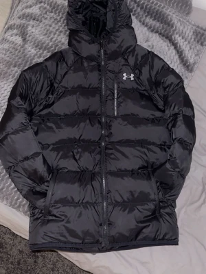 Under armour puffer jacka - Snygg svart pufferjacka från Under Armour med huva och dragkedja framtill. Jackan har en bröstficka med dragkedja och två sidofickor. Perfekt för kalla dagar med sin vadderade design och sportiga look. Köpt från jdsport för 1200kr och är använd några gånger 