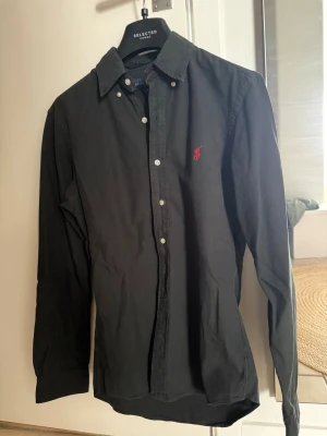 Svart skjorta från Ralph Lauren - Klassisk svart skjorta från Ralph Lauren med lång ärm och knappar framtill. Skjortan har en röd broderad logga på bröstet och är tillverkad i bomull. Perfekt för en stilren look.