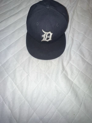 Svart New Era Detroit Tigers keps - Snygg svart keps från New Era med Detroit Tigers-logga broderad i vitt framtill. Klassisk rak skärm och ventilerande hål upptill. Kepsen är gjord i bomullstyg och har New Era-loggan broderad på sidan. Perfekt för dig som gillar sportig streetstyle.