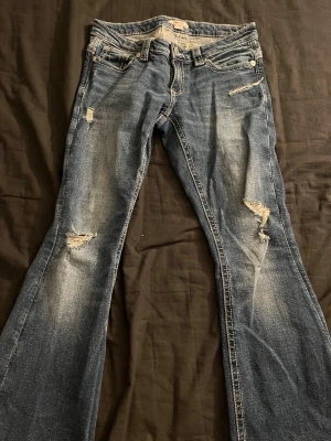 Blå bootcut jeans med slitningar - Säljer ett par blå bootcut jeans med slitningar och snyggt broderad bakficka. Jeansen har ljusa tvättade partier och detaljerade sömmar. Är från young gina och är låååg midjade.
