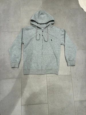 Grå zip-hoodie från Polo Ralph Lauren - Säljer en grå zip-hoodie från Polo Ralph Lauren med huva och dragsnören. Hoodien har ett litet svart logotryck på bröstet, ribbade muddar och fickor framtill. Perfekt för dig som gillar klassisk och stilren streetwear.